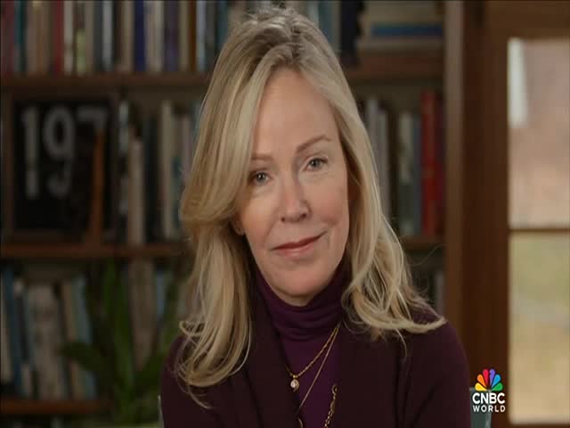 A woman with blonde hair and a burgundy turtleneck smiles, her gaze directed towards the camera. Behind her, bookshelves and the CNBC World logo indicate she's on a news broadcast.
