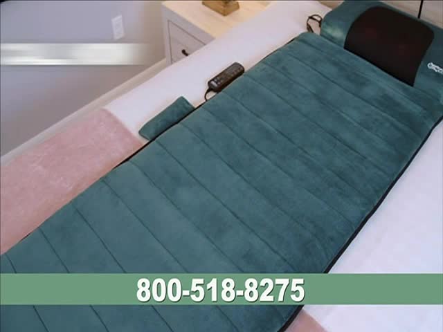 A teal massage mat with a built-in headrest lies on a white bed, a remote control resting nearby. The CNBC World graphic at the bottom displays a phone number.
