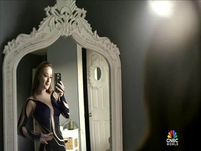 A woman in a patterned dress takes a selfie in a large mirror. The CNBC World logo is visible in the bottom right corner.
