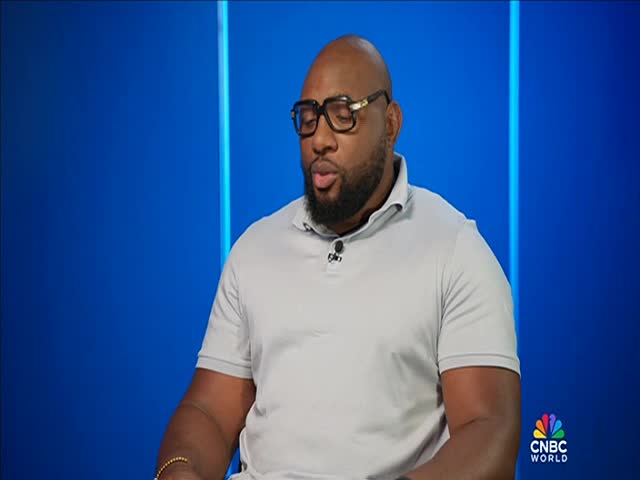 A man in a light gray polo shirt and thick glasses sits in front of a blue backdrop. He appears to be speaking, a small microphone clipped to his shirt.
