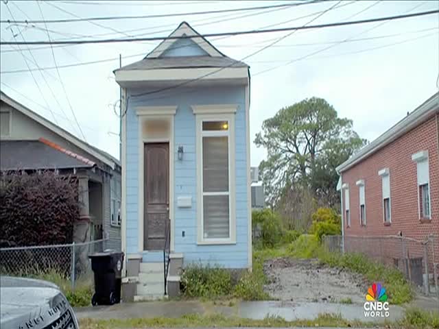 A small, light blue house stands out against the overcast sky, with a dark wooden door and a tall window.  The CNBC World logo is in the corner, and the scene is a residential street in the United States.
