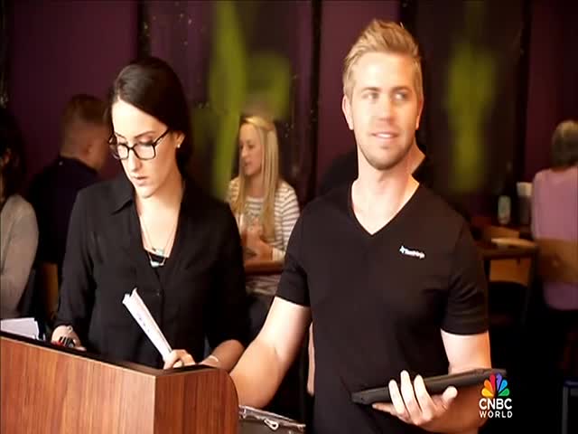 A woman in glasses, holding a stack of papers, stands behind a wooden podium.  Beside her, a man in a black shirt holds a tablet, looking toward the camera.
