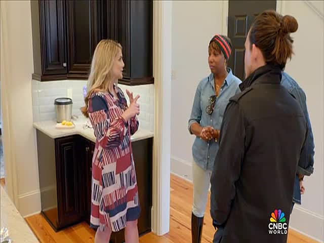 A woman in a patterned dress gestures with her hands, seemingly explaining something to two people. The three stand in a kitchen, and the CNBC World logo is visible in the corner.
