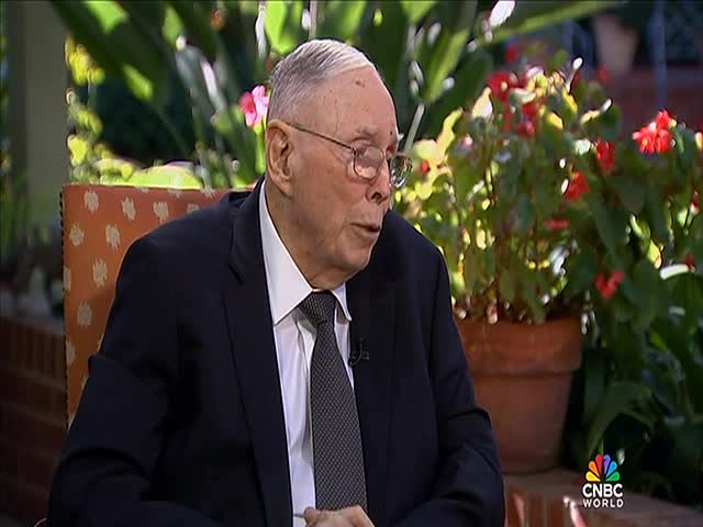 A man in a dark suit, wearing glasses, is speaking, his mouth moving as he looks off to the side.  Behind him, a potted plant with red flowers sits on what appears to be a patio, and the CNBC World logo is visible.
