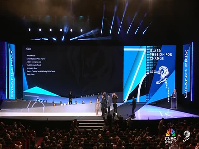 A group of people stand on a stage, some with microphones, while a man at a podium addresses the audience. The CNBC World logo is visible, and the backdrop displays the words "Glass: The Lion for Change."

