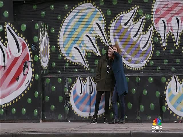Two women are posing for a selfie in front of a wall covered in colorful graffiti. One points her phone towards the camera while the other smiles, their image framed by the artwork.
