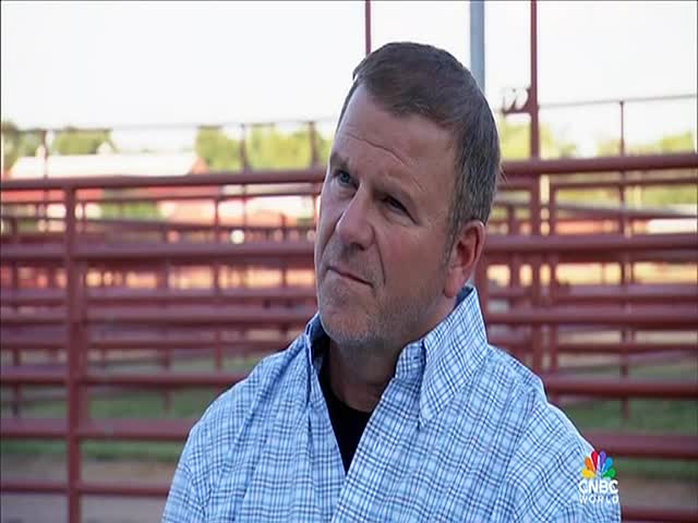 A man in a blue-and-white checkered shirt looks off to the side, with a red metal fence behind him. The CNBC World logo is visible in the corner.
