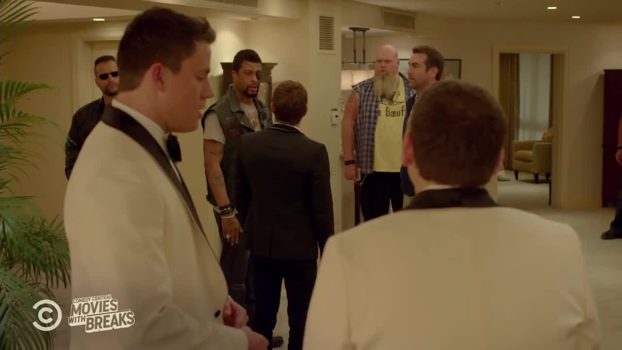 A group of men in suits and leather vests are gathered in a hotel hallway. Two men in white tuxedos stand facing the others, who seem to be blocking the way.
