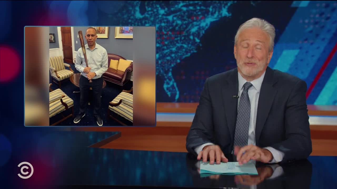On the Comedy Central set, Jon Stewart sits at a desk, looking at the camera. A photo of a man holding a baseball bat in what looks like a government office is displayed in a box next to him.
