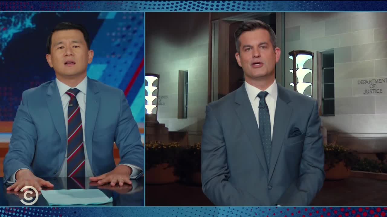 Two men in suits are on a split screen, both speaking on a Comedy Central show. One is in a studio with a blue backdrop, while the other stands outside the Department of Justice building.
