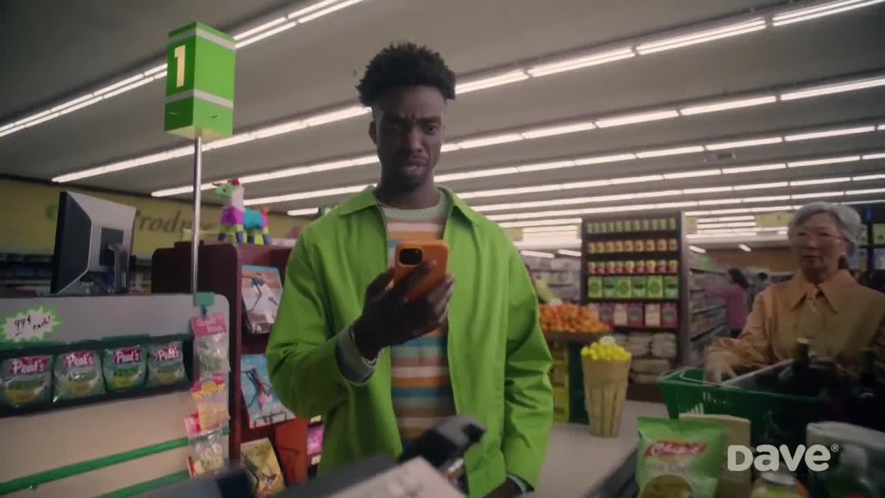 A man in a bright green jacket stares intently at his phone, his brow furrowed. Behind him, a woman in a yellow shirt waits patiently at the checkout counter.
