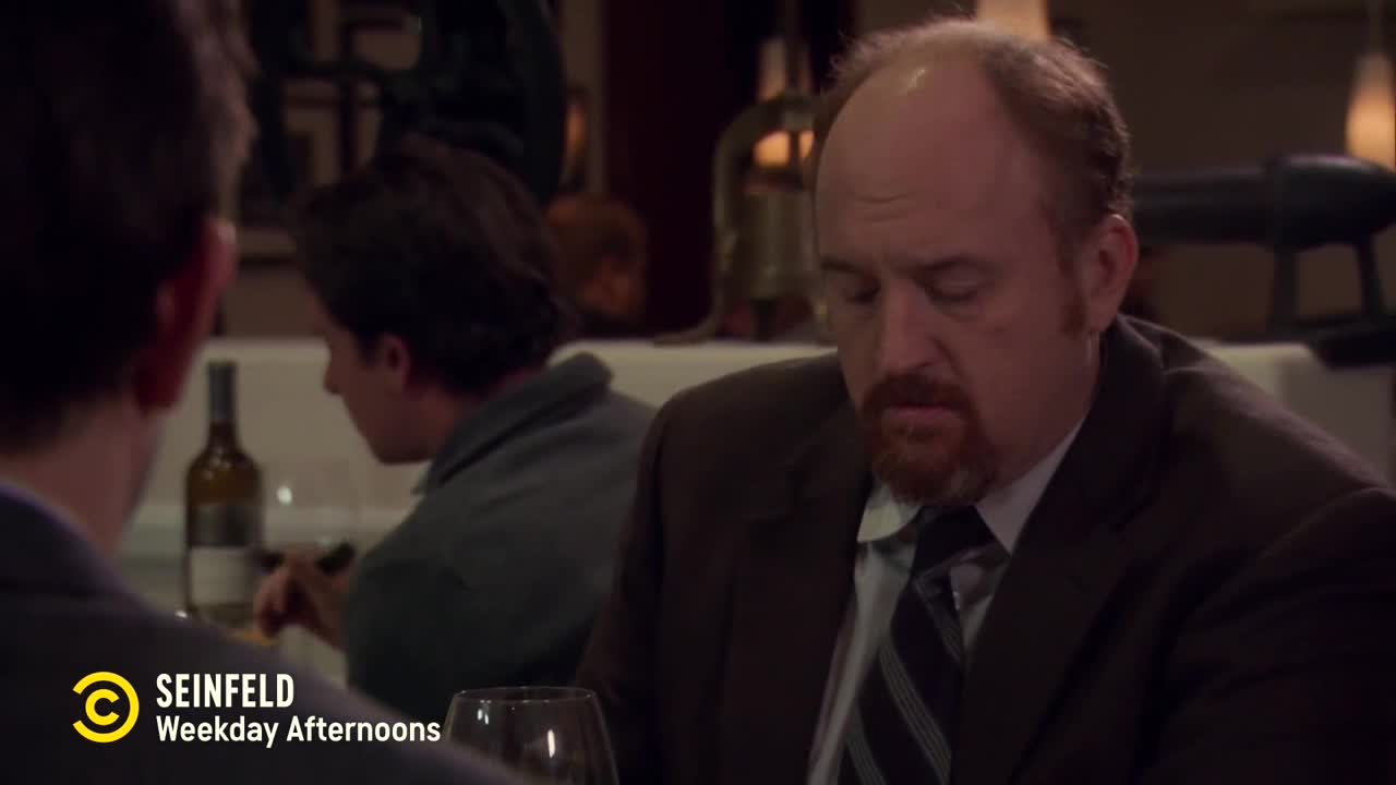 A man in a suit and tie stares down at the table, his face somber. Across from him, another man is partially visible, and a bottle of wine sits between them.
