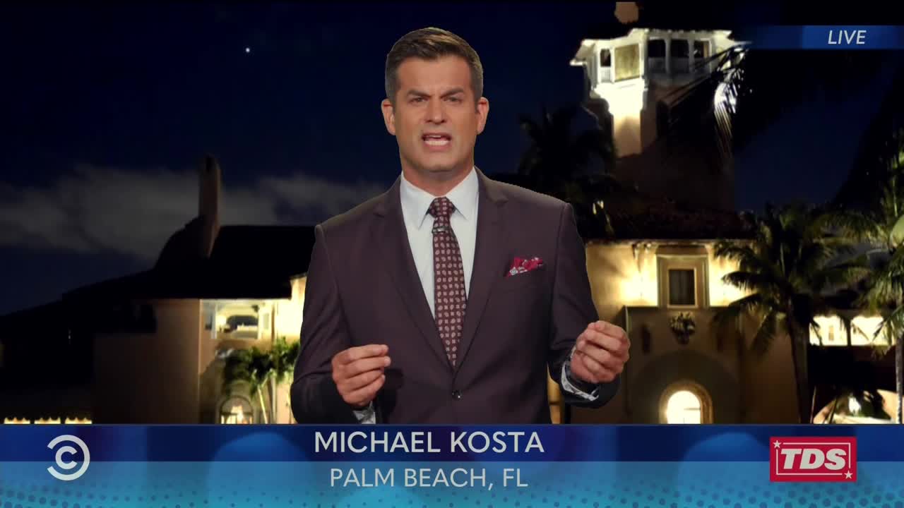 Michael Kosta, in a suit, gestures emphatically as he speaks, the backdrop a stately building lit up at night. The Comedy Central logo and location are displayed at the bottom of the screen.
