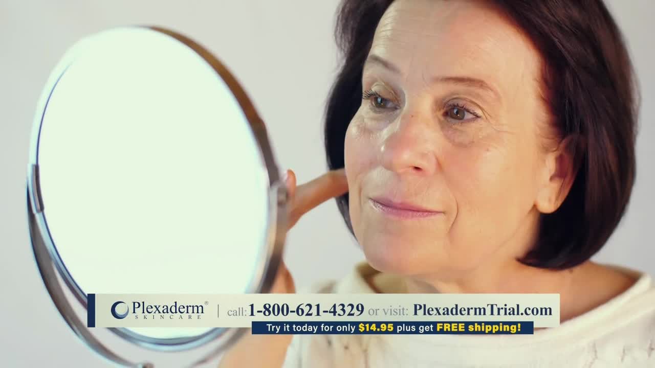 A woman is holding a round mirror, studying her face closely. Her finger gently touches her cheek as she looks for something in the reflection.
