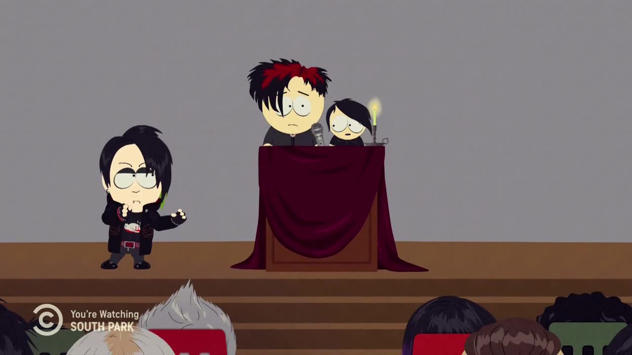 A kid dressed in black stands to the left, gesturing with his hand. Two more, looking nervous, stand behind a draped podium, with a candle burning on a small stand.
