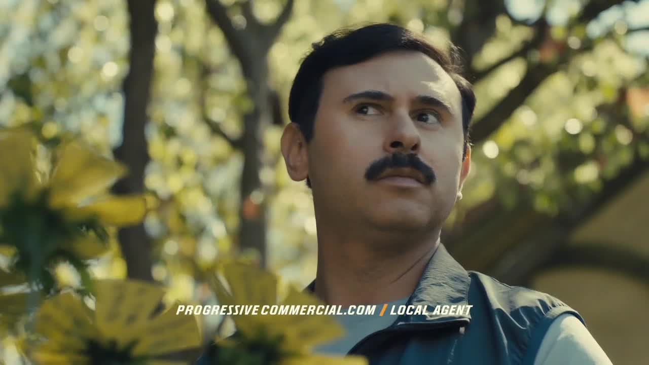 The man with the mustache is looking off to the side, framed by yellow flowers. The background is a blur of green and brown, and a website address is visible at the bottom.
