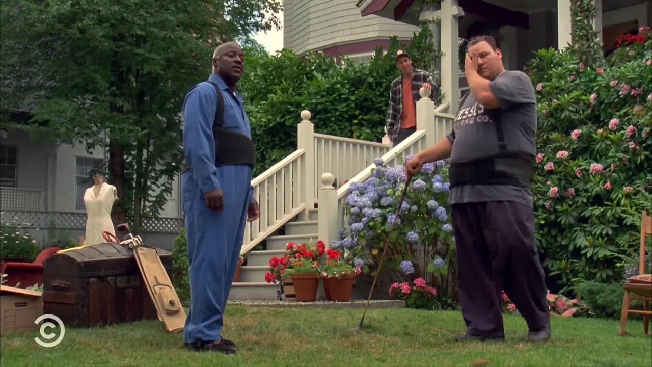 A man in a blue jumpsuit stands on the lawn, watching as another man, wearing a back brace, gestures with a golf club. A third man leans on the porch behind them, looking on.
