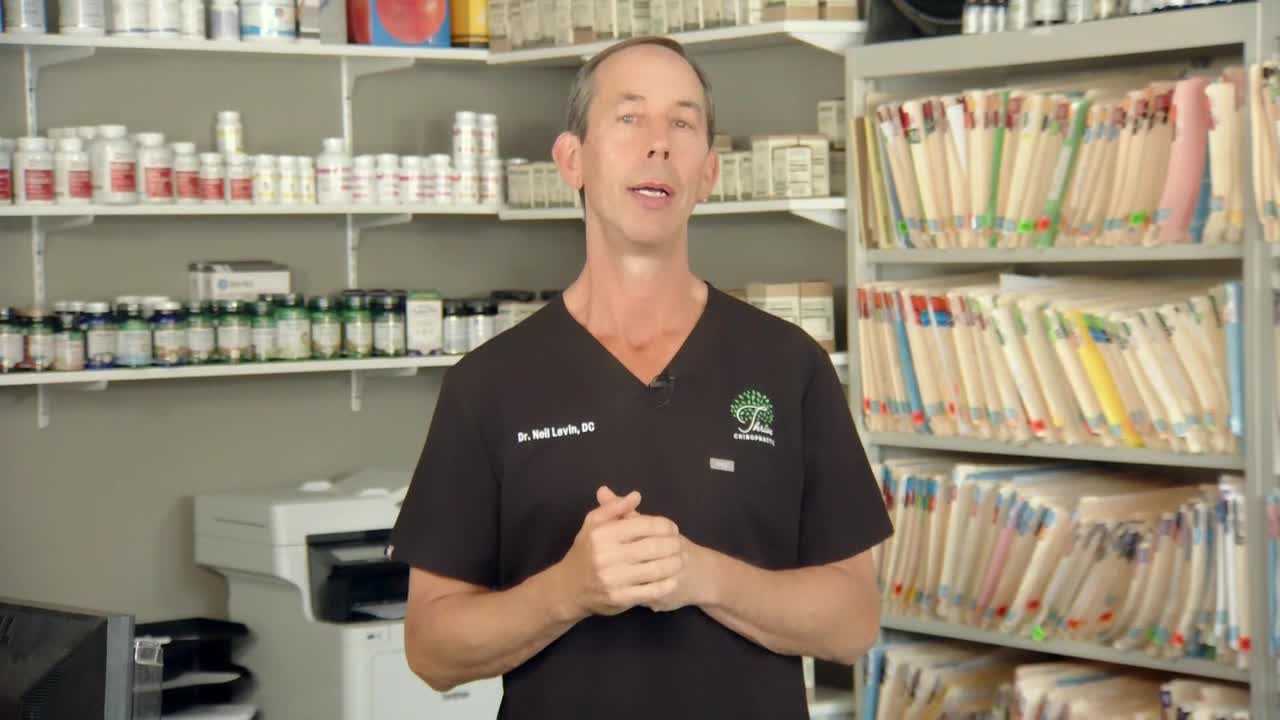 A man in a black scrub top is talking, his hands clasped in front of him. Behind him, shelves are filled with supplements and file folders.
