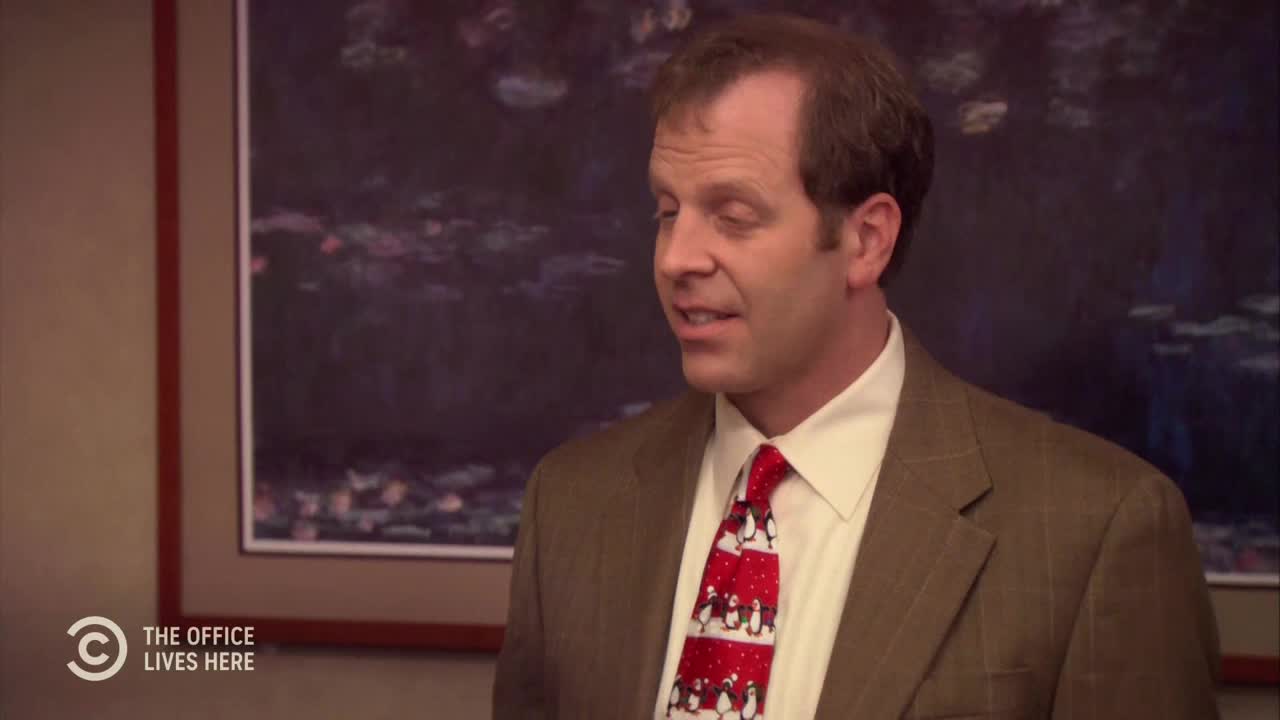 A man in a brown suit and a penguin-patterned tie is speaking, his mouth slightly open. The Comedy Central logo is visible in the corner, and a framed picture hangs behind him.
