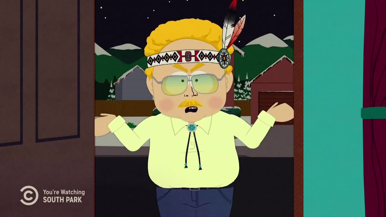 A man in a Native American headdress stands in a doorway, arms raised in a gesture of confusion. His face is scrunched up, and he looks like he's about to say something.

