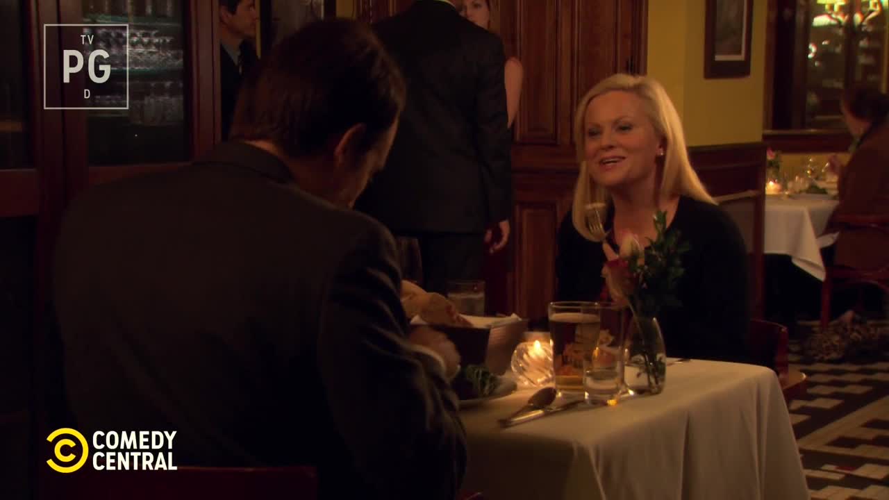 A woman with blonde hair, holding a wine glass, smiles at someone off-screen. Across the table, a man in a suit is focused on his meal in a dimly lit restaurant.
