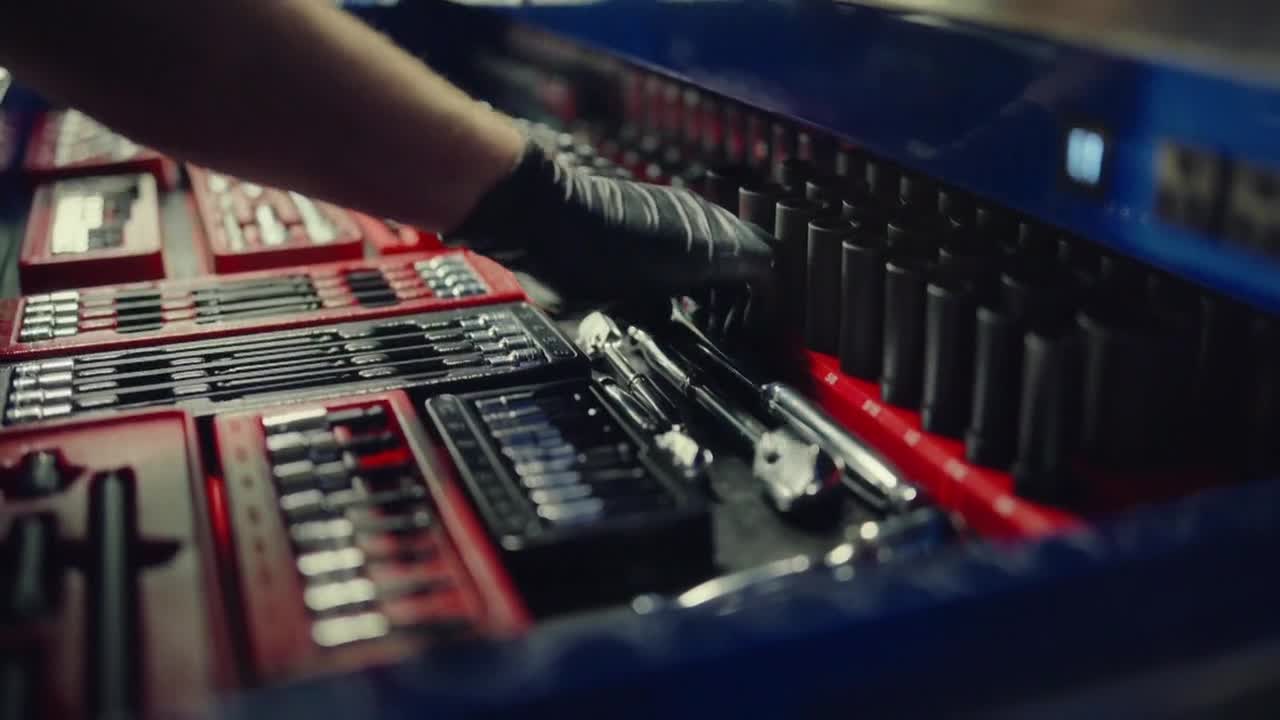 A gloved hand is reaching into a drawer filled with tools, carefully selecting a socket. The drawer is organized with red trays and neatly arranged tools, ready for use.
