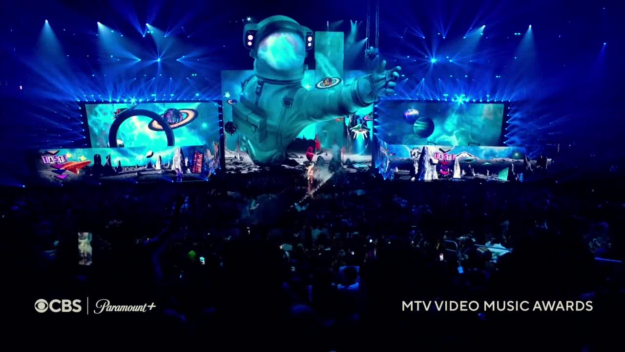 A giant astronaut, rendered in blue, dominates the stage backdrop, its hand outstretched. Below, performers move in front of a large screen depicting planets and space, bathed in bright spotlights.
