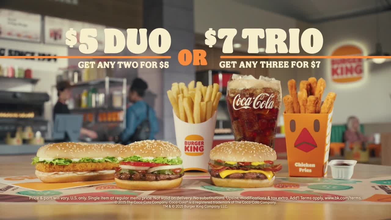 A spread of Burger King food sits on a counter, with options for a $5 Duo or a $7 Trio. Behind the food, two people are visible, likely employees, in what looks like a quick-service restaurant.
