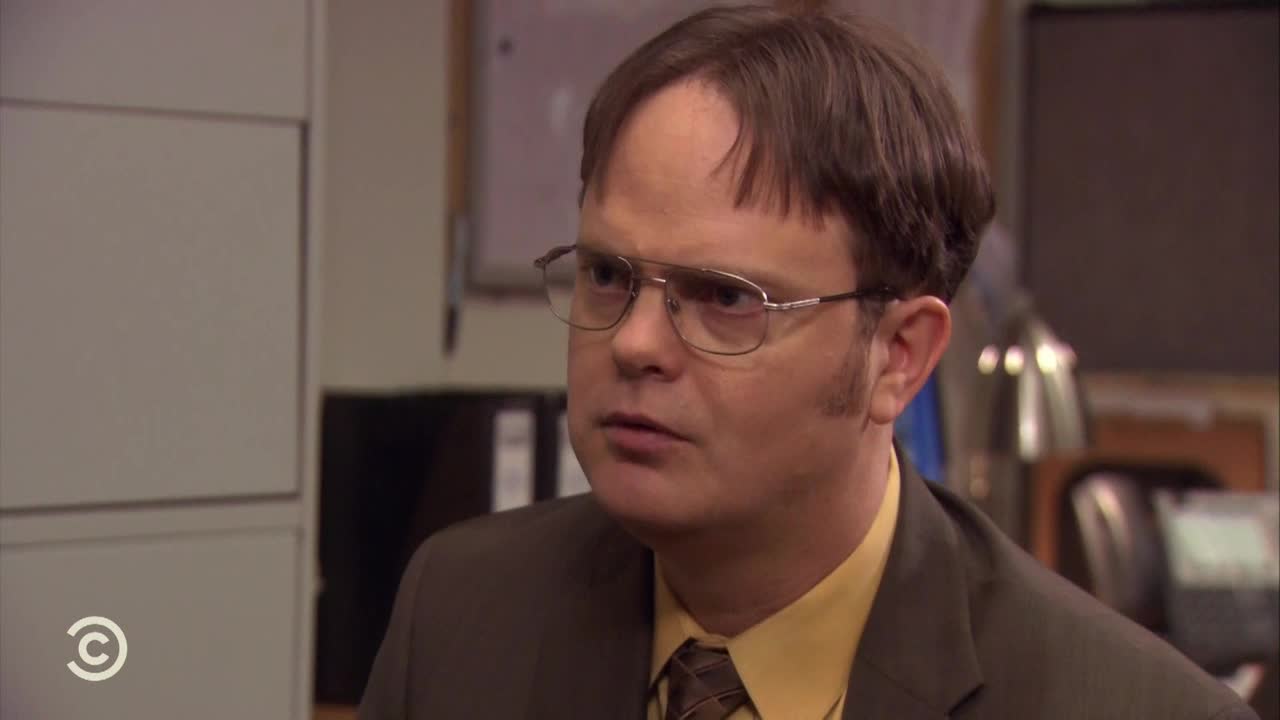Dwight Schrute, from the United States, is staring intently, his brow furrowed. He's wearing a brown suit, a yellow shirt, and those distinctive glasses.

