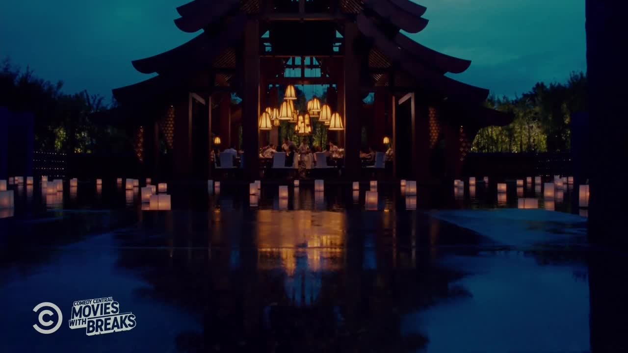 The scene is lit by the warm glow of candles floating on the water and hanging from the ceiling of a large wooden structure. People are dining at tables inside, their reflections shimmering in the wet surface.

