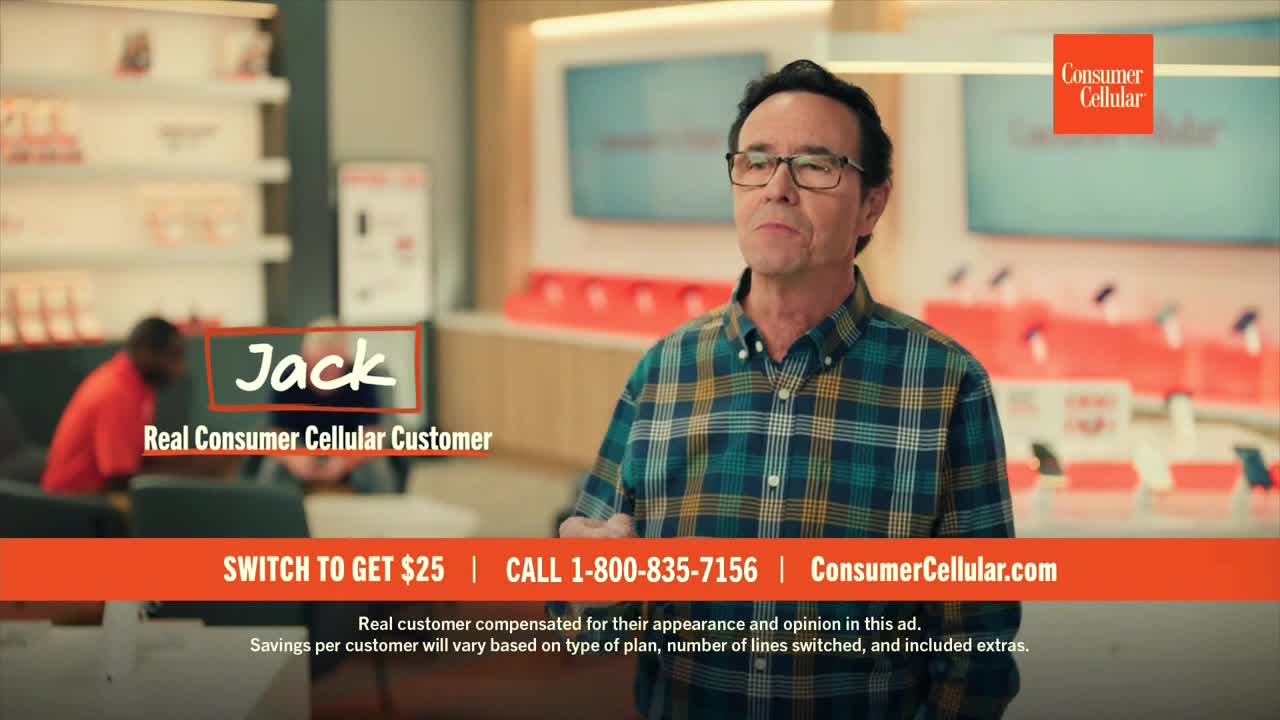 A man in a plaid shirt stands in what looks like a phone store, gesturing with his hands. The screen displays the Consumer Cellular logo and a call to action offering $25 to switch.
