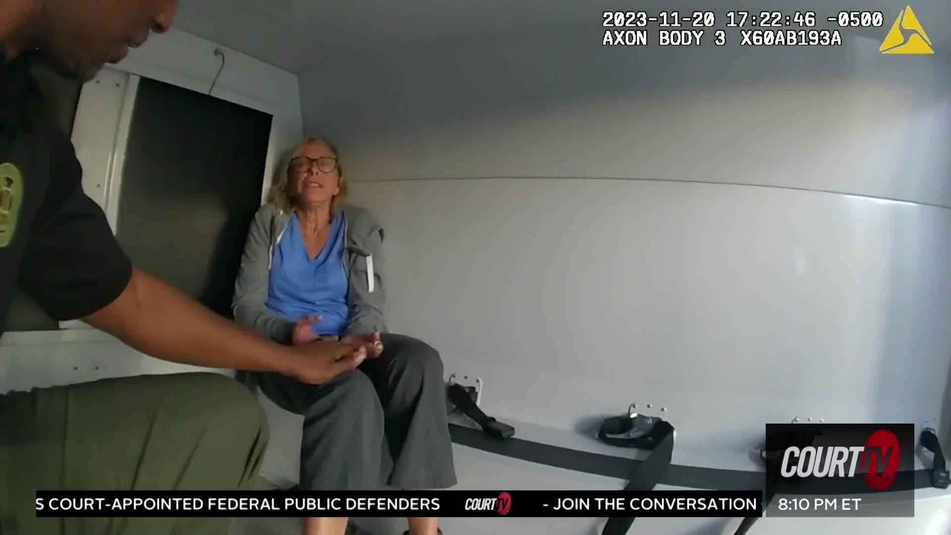 A woman in a blue shirt sits in the back of a vehicle, her hands held out. A person in a dark uniform is reaching toward her, their hand outstretched.
