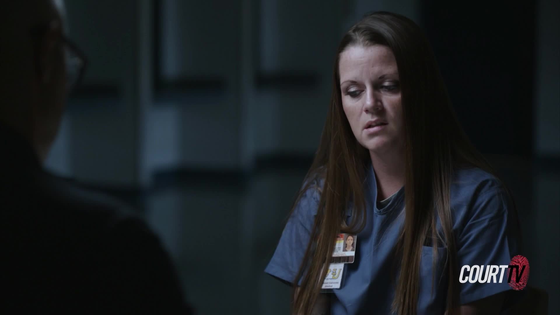 A woman with long brown hair sits at a table, looking down with a troubled expression. She's wearing a blue uniform, and the Court TV logo is visible in the corner.
