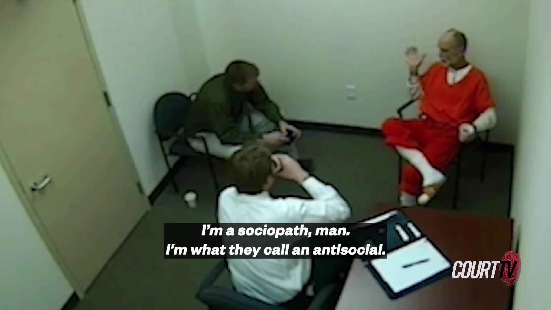 A man in an orange jumpsuit sits in a chair, gesturing with his hand while speaking. Two other men are in the room, one on the phone and the other seated and leaning forward.
