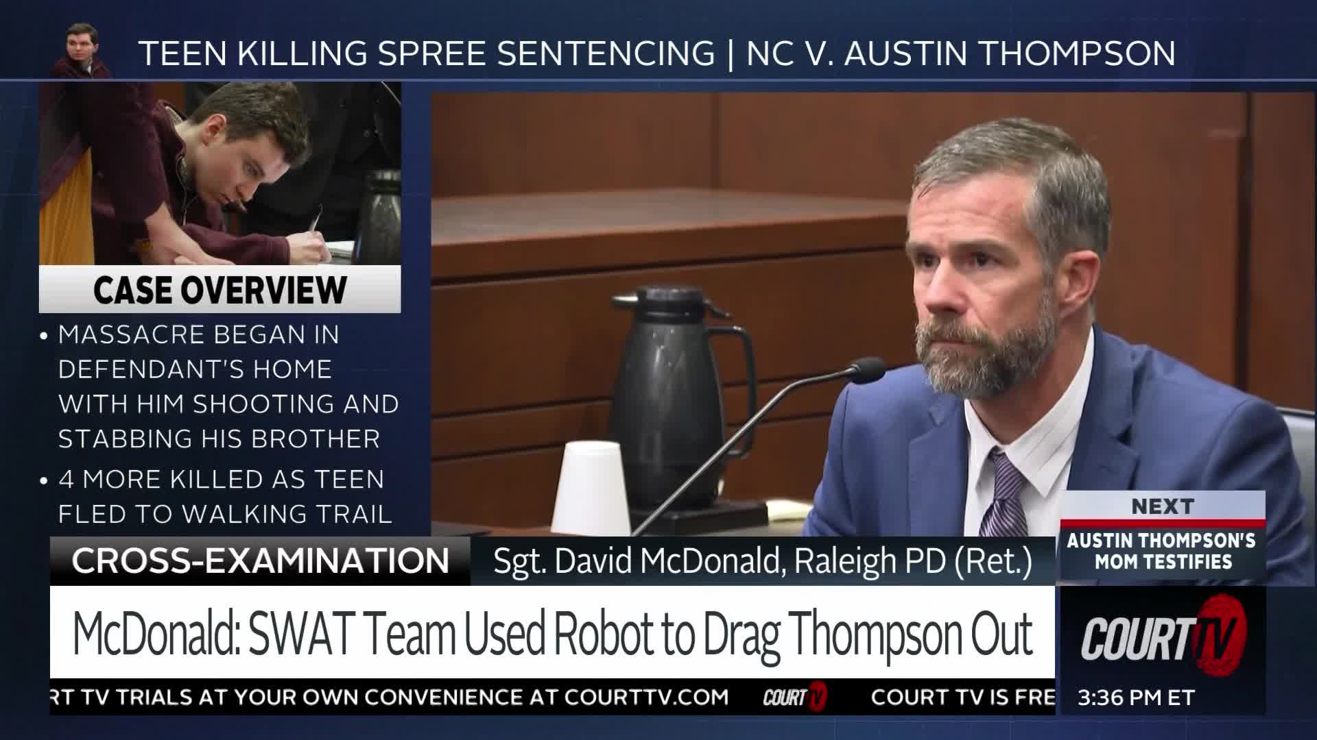 A man in a blue suit with a beard sits at a table, speaking into a microphone. On a screen to his left, a young man in a maroon shirt bends over a desk, looking down at papers.