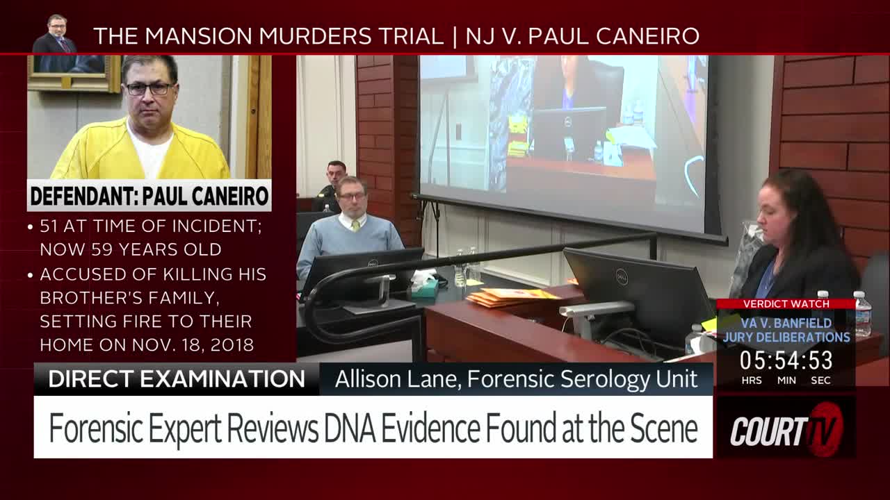 A woman in a dark jacket sits at a table, looking at a laptop. On a large screen behind her, a man in a suit stands at a podium.