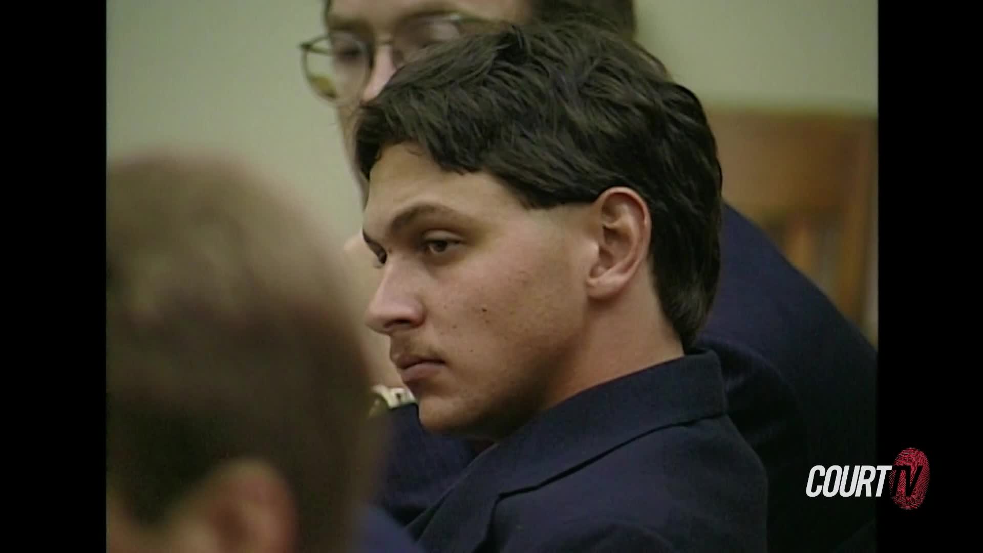 The young man in the dark suit turns his head slightly, his gaze fixed on something just out of frame. Another person, wearing glasses, is visible behind him, their face blurred.