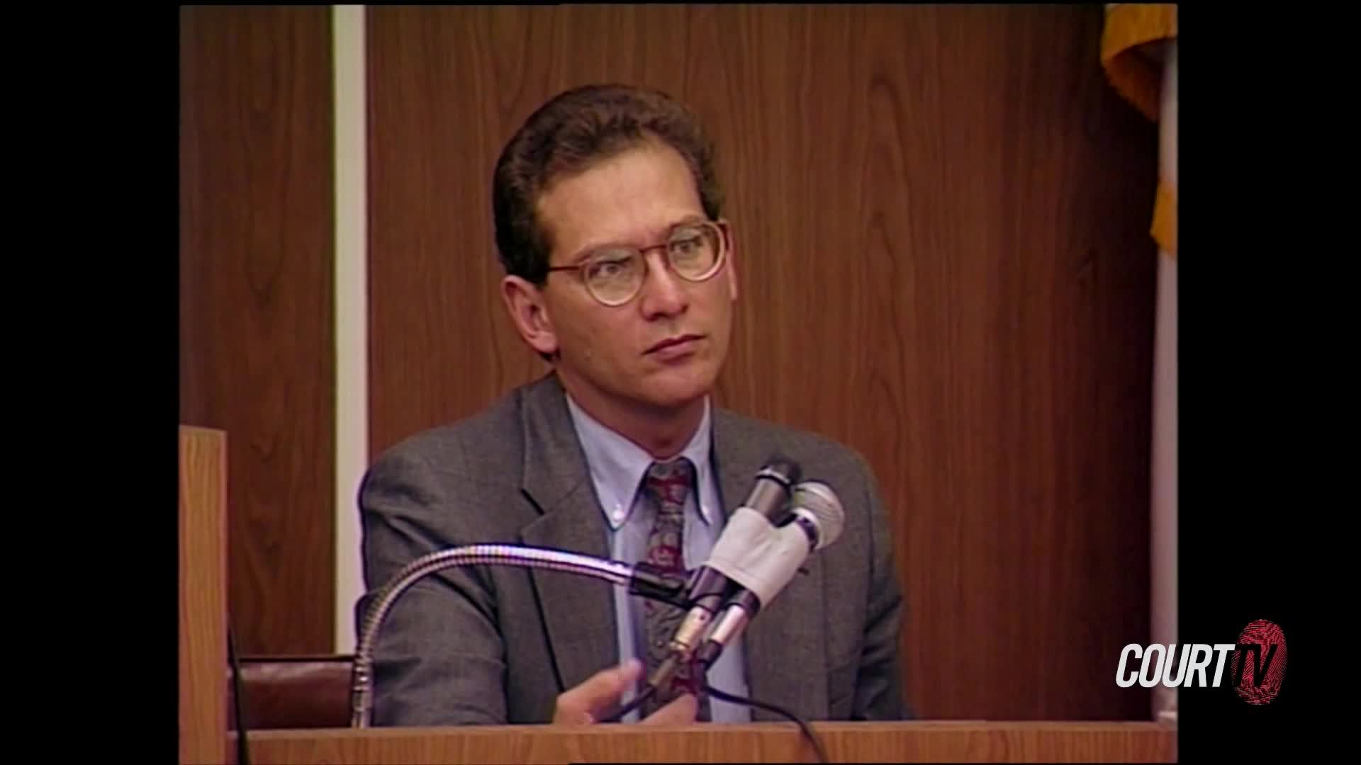 A man in a suit and glasses sits at a wooden desk, looking intently to his left. Two microphones stand before him, their silver necks curving upwards.