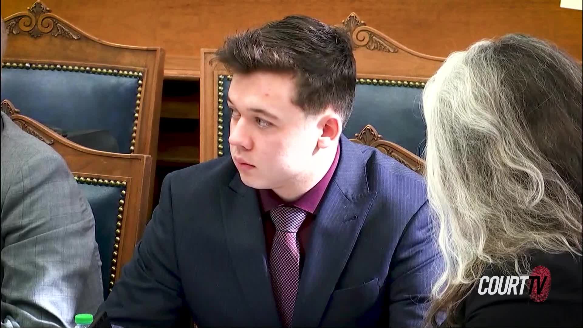 He turns his head, his gaze fixed on something off-camera. The courtroom's polished wood and dark upholstery surround him.