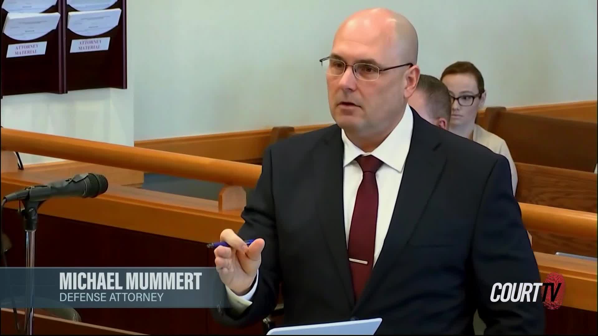The defense attorney gestures with a pen as he speaks. Behind him, a woman in glasses sits in the jury box.