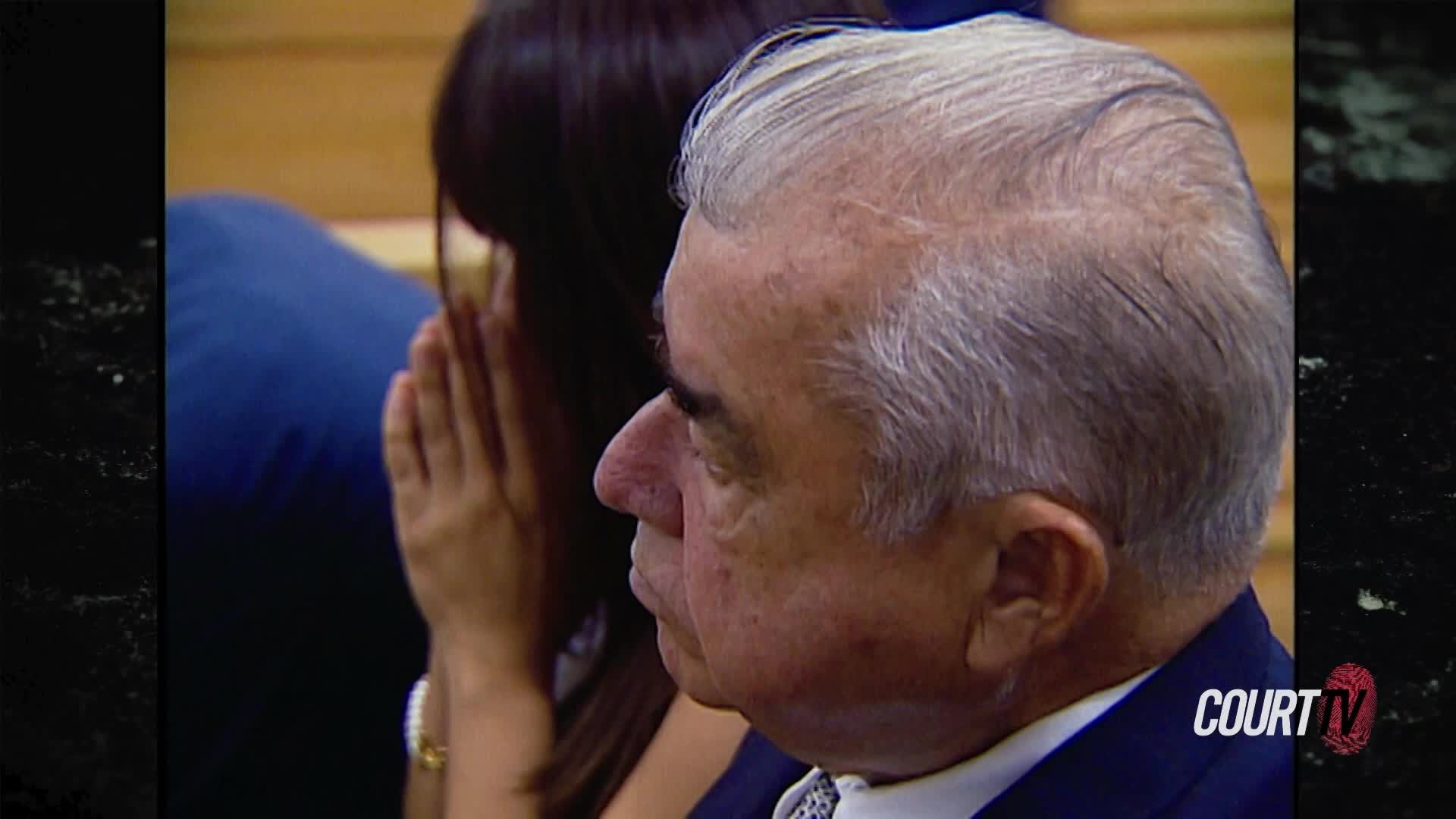 The man in the suit, his profile sharp against the wooden benches, turns his head slightly. Beside him, a woman covers her face with her hands, her shoulders shaking.