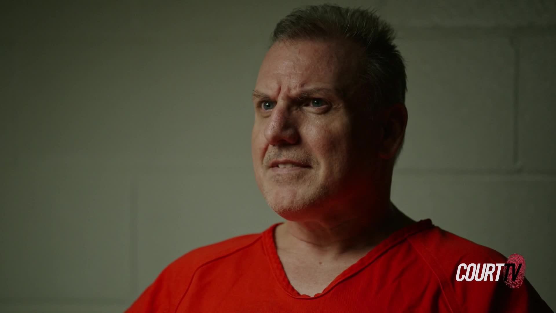 A man in an orange jumpsuit stares intently, his brow furrowed. The harsh lighting casts deep shadows across his face, highlighting his worried expression.