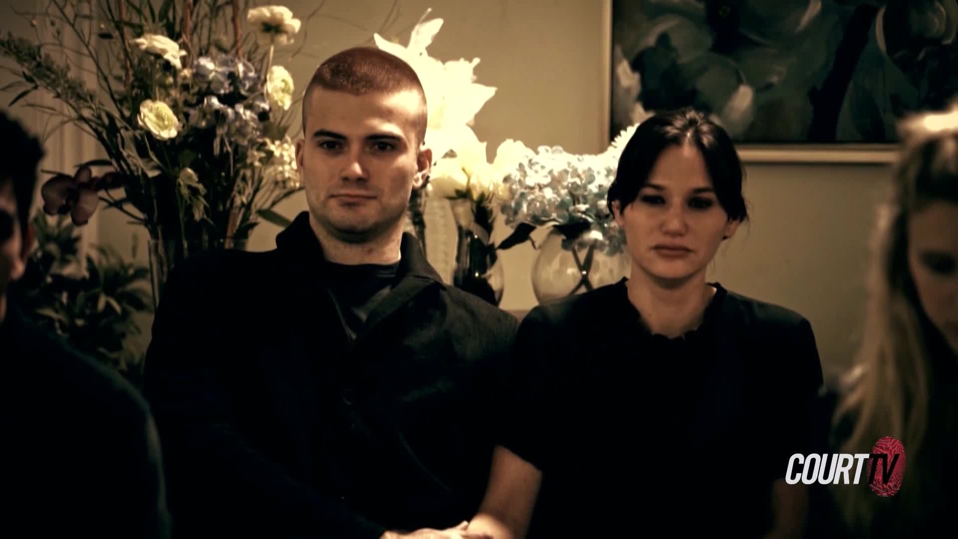 A young man with a close-cropped haircut stares directly ahead, his expression unreadable. Beside him, a woman with dark hair looks down, her face etched with a somber mood.