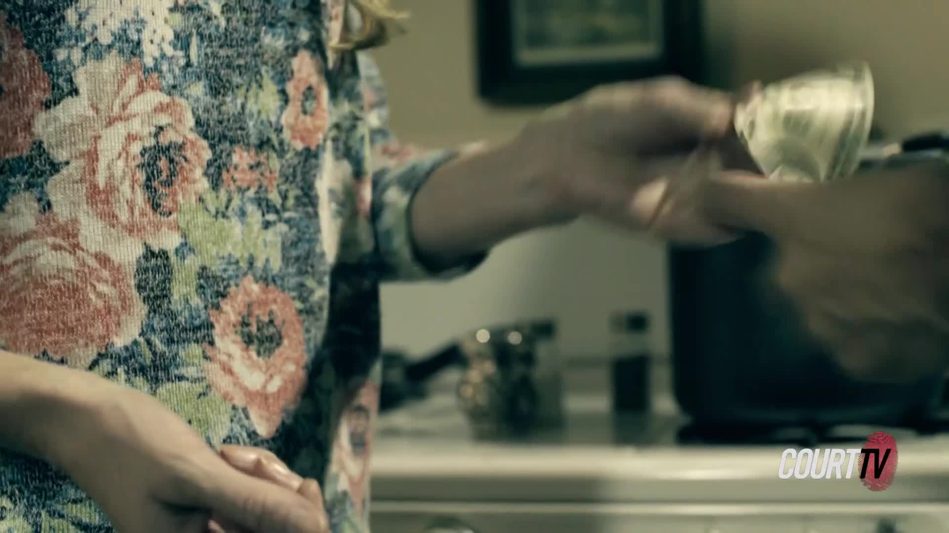 A woman in a floral blouse offers a small glass to another person. The glass appears to hold a liquid, and the exchange happens in a kitchen setting, perhaps during a moment relevant to a Court TV case.