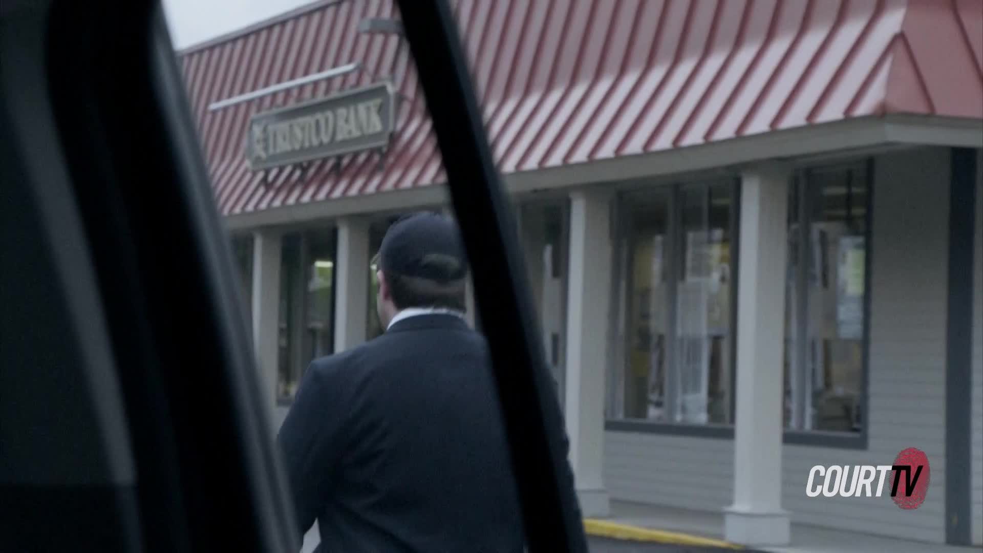 A man in a dark suit and baseball cap walks away from a car towards a Trustco Bank. The red and white striped awning of the bank is visible overhead.