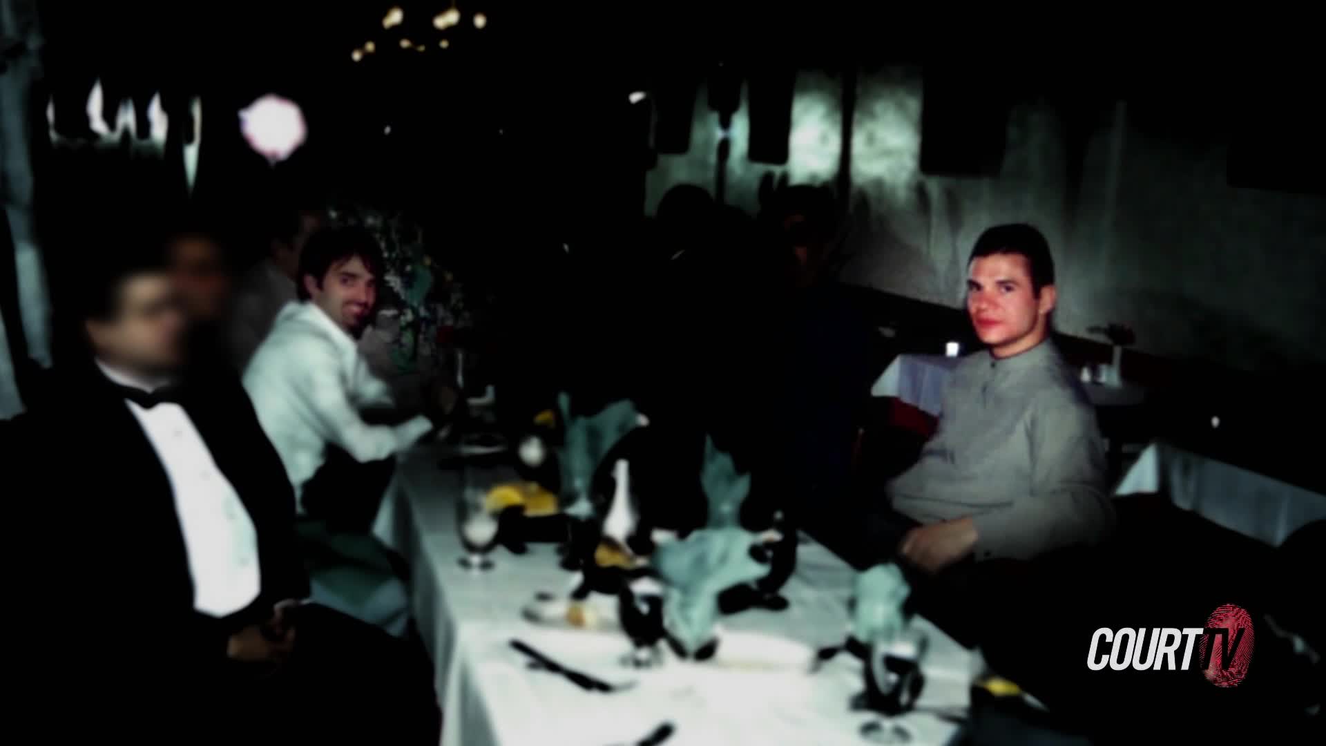 A man in a light-colored shirt sits at a table in a dimly lit room, looking directly at the camera. Across the table, another man in a white shirt smiles, his hand resting on the table.