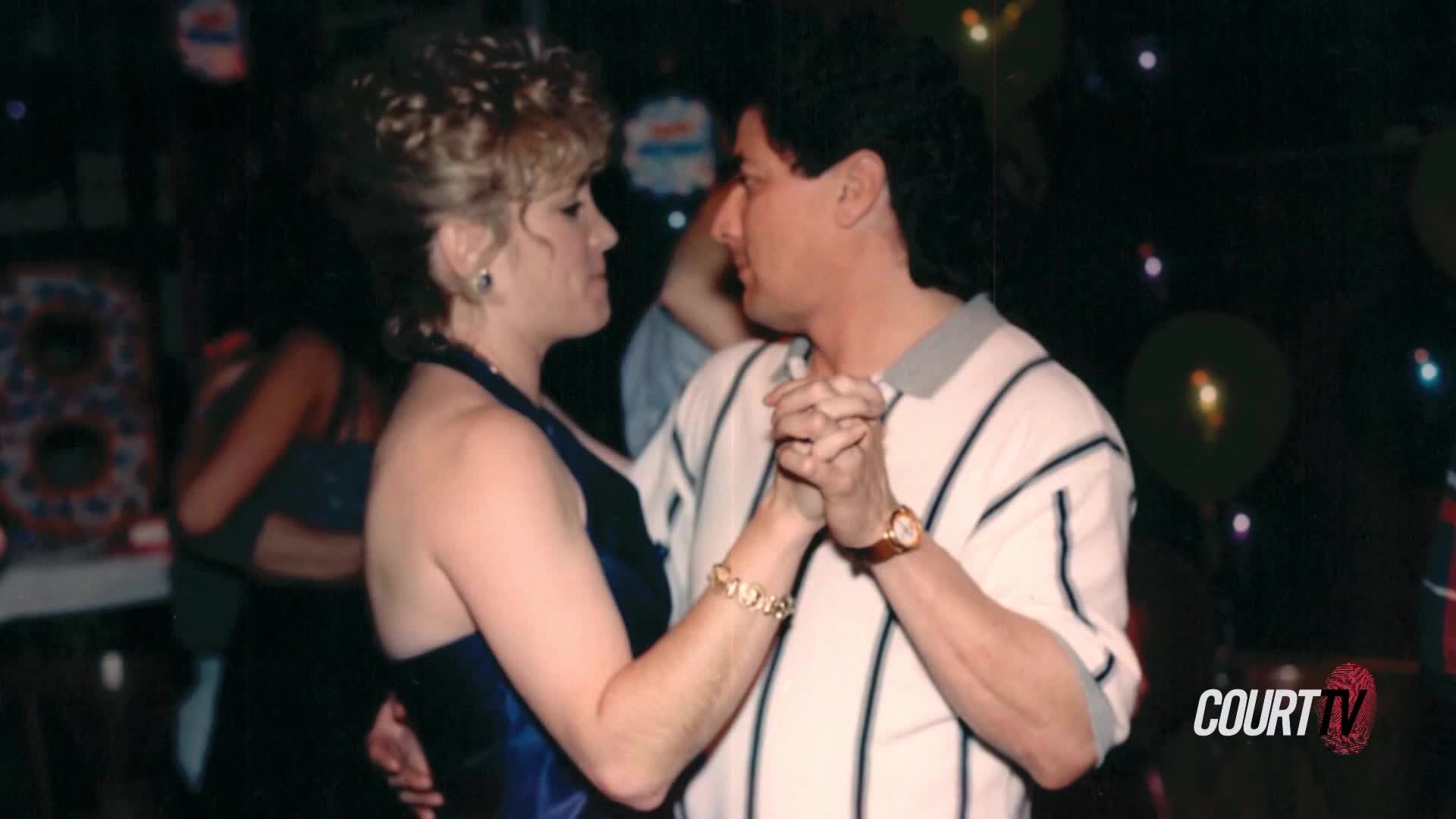 A couple dances closely, their hands clasped. The woman wears a dark blue dress, while the man sports a striped polo shirt.