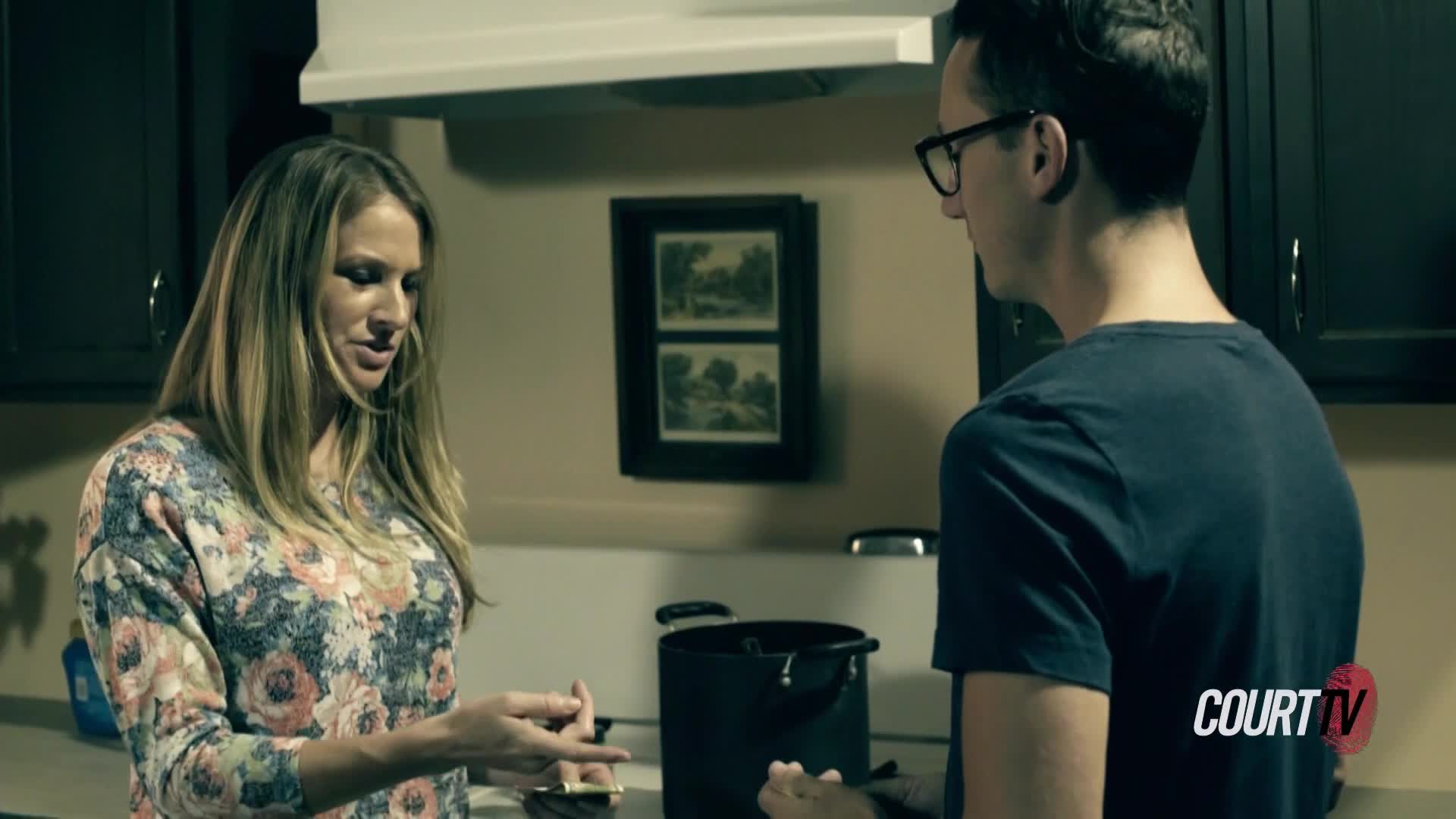 A woman in a floral shirt gestures with her hands, speaking to a man in a dark t-shirt and glasses. They stand in a kitchen, a large black pot between them.