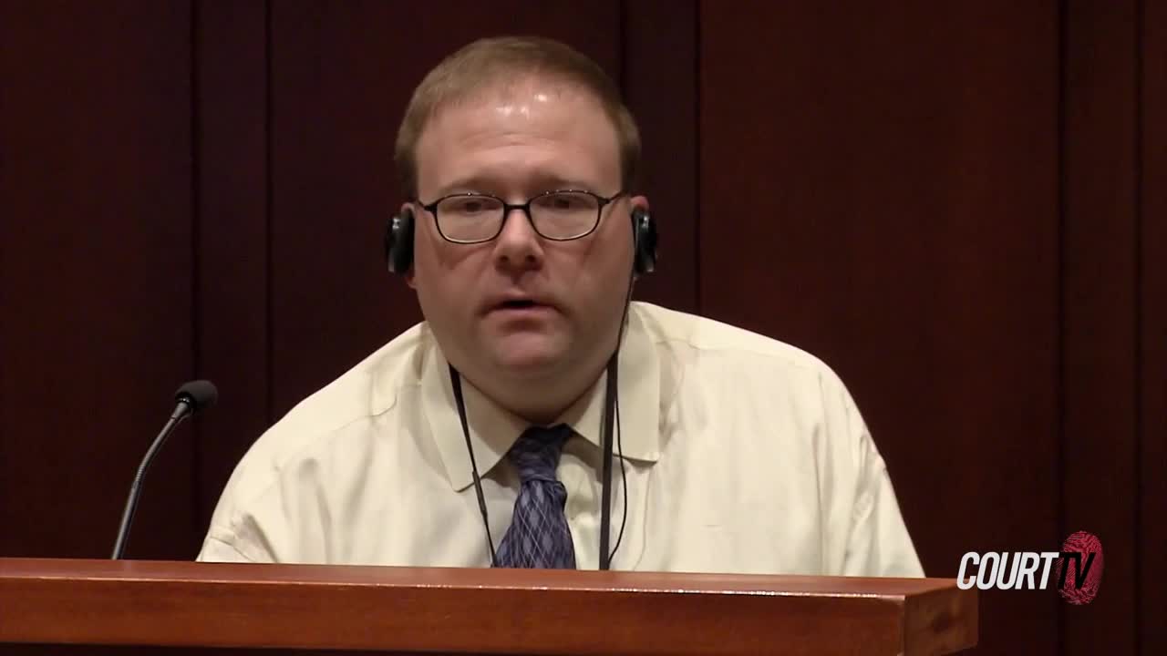 A man with glasses and headphones speaks into a microphone. He's wearing a light-colored shirt and a patterned tie, seated behind a wooden railing.