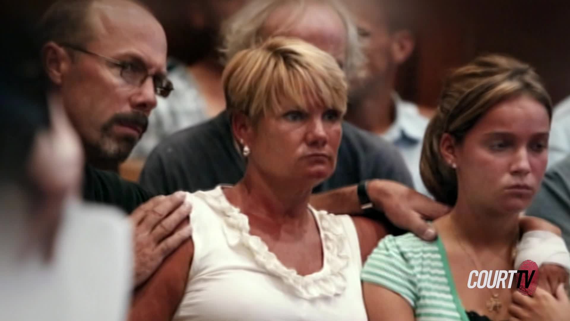 A man places his hand on a woman's shoulder, his gaze fixed forward. Beside her, a younger woman looks down, her arm bandaged.